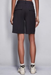 Pantaloni shorts neri su misura con lunghezza a metà coscia, due ampie tasche posteriori e dettaglio del logo in rilievo, abbinati a sneakers bianche alte.