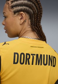 Maglia gialla del Borussia Dortmund con accenti neri. Presenta un grande testo "DORTMUND" sulla schiena e il logo Puma sulla spalla. Tessuto testurizzato.
