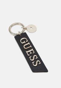 Czarny skórzany brelok do kluczy z wytłoczonym złotym napisem "GUESS", z srebrnym pierścieniem kluczowym i okrągłą metalową zawieszką z wycięciem w kształcie trójkąta.