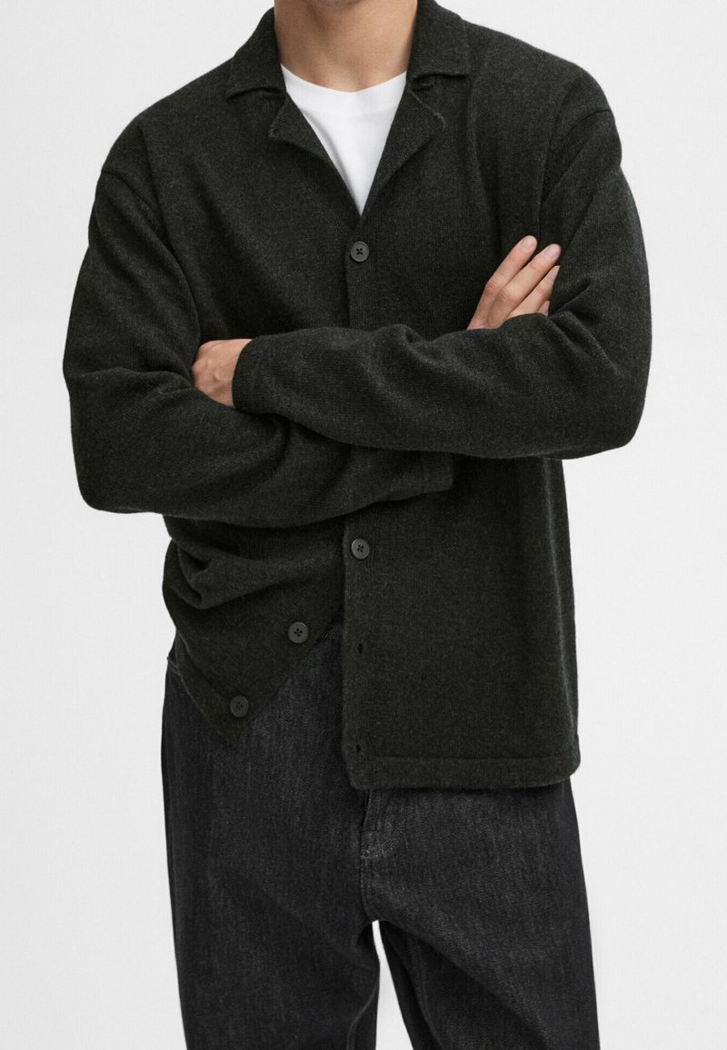 Homme portant un cardigan en laine noire boutonné sur une chemise blanche et un pantalon noir, bras croisés devant un fond uni.