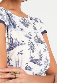 Kortärmad topp med vit bas som har blå blommönster och landskapsmönster; marinblå kant vid ärmarna. Guldringdetalj på handen.