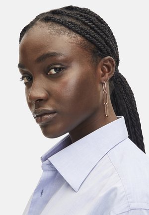 Long, thin gold earrings with oval links. The model wears a light blue shirt with a pointed collar, featuring a smooth fabric texture.