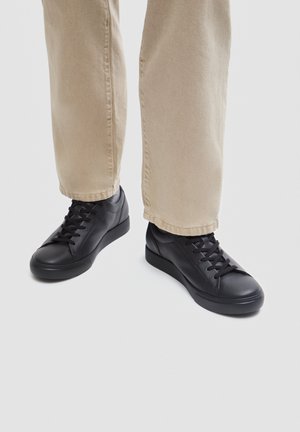 Persona che indossa pantaloni beige e sneakers nere in pelle con lacci, in piedi su uno sfondo grigio chiaro.