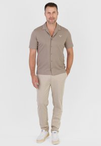 Homme portant une chemise taupe à manches courtes et à boutons, un pantalon beige et des baskets blanches, debout avec une main dans la poche sur un fond gris uni.