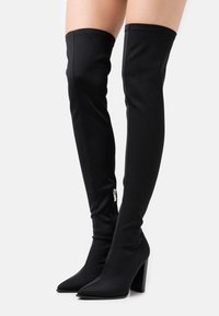 Bottes noires au-dessus du genou en tissu extensible, avec un bout pointu, un talon bloc et une fermeture éclair latérale pour un enfilage facile.