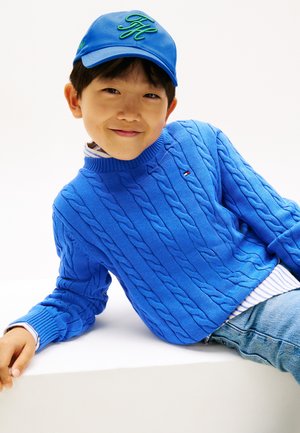 Băiat zâmbitor îmbrăcat într-un pulover albastru cu model împletit, șapcă albastră cu litere verzi, cămașă în dungi și blugi, sprijinit de o suprafață albă.