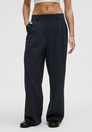 Frau trägt hoch taillierte, breitbeinige schwarze Hose mit Gürtelschlaufen, kombiniert mit schwarzen Schuhen und einem kurzen weißen Top, steht vor einem einfarbigen Hintergrund.