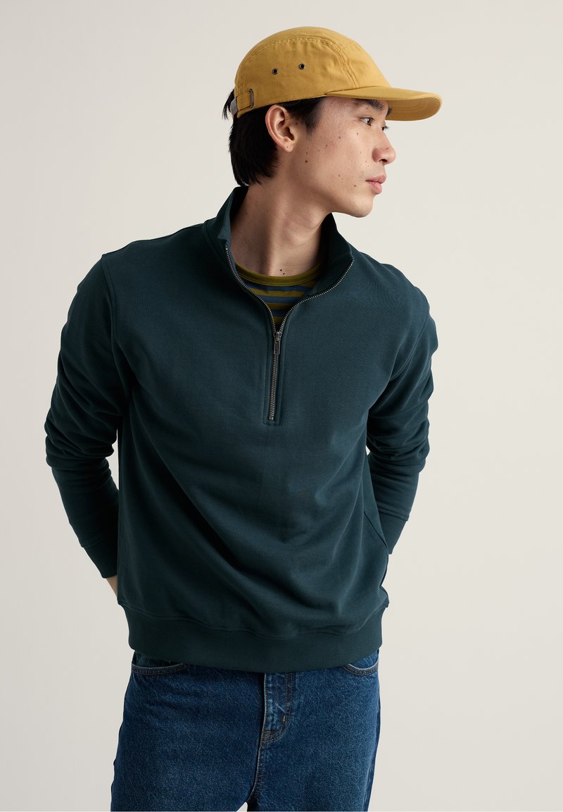 Homme portant une casquette jaune moutarde, un sweat-shirt à fermeture éclair quart vert foncé, et un jean bleu, regardant vers sa droite avec les mains derrière le dos.