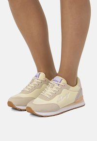Zapatillas deportivas de color amarillo claro con detalles en beige, material sintético, diseño con cordones, superposiciones texturizadas y suelas de goma con dibujo de caucho.