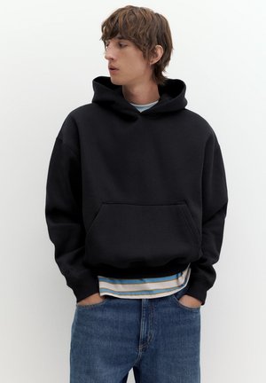 Un jeune homme portant un sweat à capuche noir et un jean bleu se tient debout, les mains dans les poches, en regardant sur le côté.