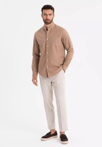 Chemise brune à manches longues avec col mandarin, poche poitrine unique, associée à un pantalon clair et des mocassins foncés.
