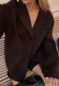 Črn blazer iz volnene mešanice, z zmanjšanim ovratnikom, sprednjimi žepi in dvema črnima gumboma; gladka tekstura in sproščen kroj.