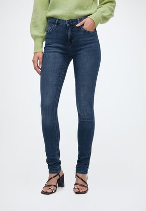 Person trägt dunkelblaue Skinny-Jeans, grünen Pullover und schwarze Riemchen-Sandalen mit Absatz, steht mit einer Hand in der Tasche vor weißem Hintergrund.