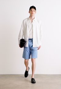 Hvit skjorte over en hvit t-skjorte, lyseblå shorts med logo, og svarte slip-on sko. Holder en svart veske.