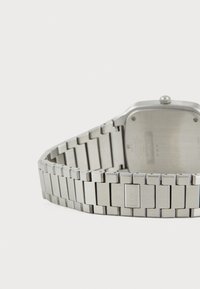 Montre en acier inoxydable avec un boîtier rectangulaire, finition brossée et bracelet en maillons. Présente un dos texturé et une gravure de marque discrète.