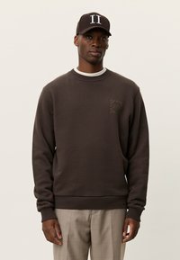 Καφέ φούτερ με ριμπ κολάρο, που φέρει ένα μικρό, τόνου με τόνο τυπωμένο λογότυπο. Φοράται με ανοιχτόχρωμο παντελόνι καρό και σκουρόχρωμο καπέλο.