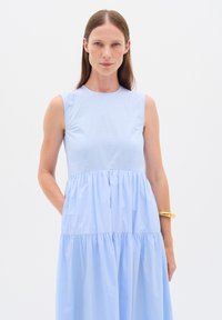 Donna con lunghi capelli castani che indossa un vestito azzurro chiaro senza maniche a balze, in piedi davanti a uno sfondo bianco con un braccialetto d'oro al polso.