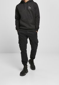 Felpa nera con tasca frontale e design grafico, abbinata a pantaloni cargo neri con tasche laterali, indossata con sneakers nere.