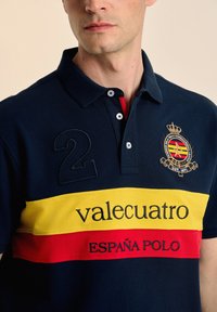 Camisa polo de color navy con rayas horizontales amarillas y rojas. Presenta un logo bordado, el número "2" y cuello con botones. Tela de algodón.