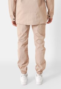 Beige cargobroek met elastische boorden, voorzien van zijzakken en een relaxte pasvorm. Draagt bij witte schoenen met zichtbare hakbranding.
