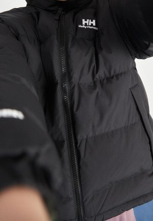 Chaqueta acolchada negra Helly Hansen con cremallera y logotipo en el pecho, vestida por una persona que extiende el brazo hacia adelante.