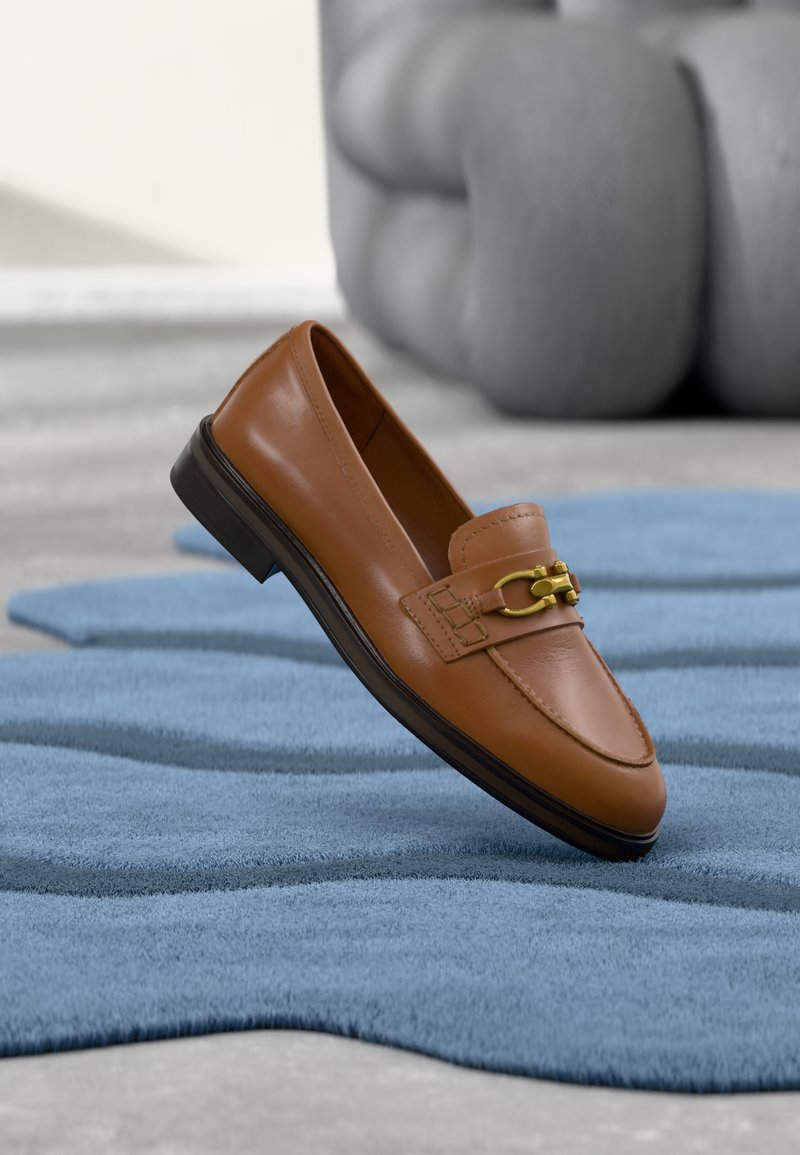Mocassins en cuir marron avec un accent de chaîne dorée, bout arrondi et semelle basse noire, posés sur un tapis texturé bleu.