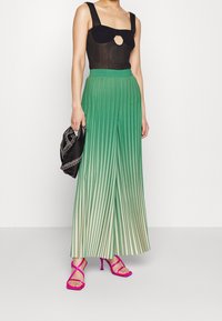 Frau in einem schwarzen ärmellosen Oberteil, grünen plissierten weit geschnittenen Hosen, die eine schwarze Nieten-Clutch und rosa Riemchen-High-Heels trägt.