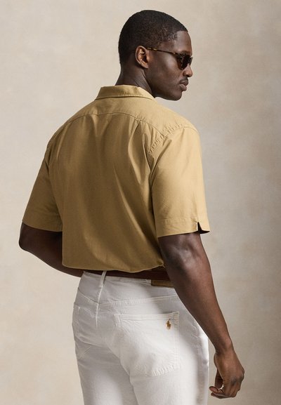 Mann mit Sonnenbrille, beige kurzärmligem Hemd und weißen Hosen, seitlich stehend vor einem einfarbigen beigen Hintergrund.