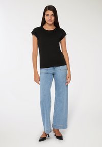Kobieta w jasnoniebieskich szerokich dżinsach, czarnym topie z krótkim rękawem i czarnych szpilkach na białym tle.