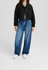 Giacca corta nera con due tasche frontali, abbinata a jeans di denim blu a vita alta e gamba larga e scarpe da ginnastica bianche. Camicia a righe sotto.