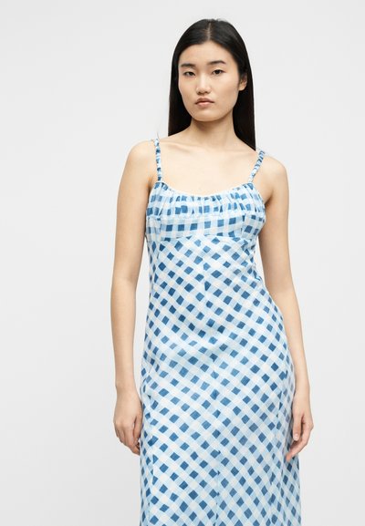 Γυναίκα που φοράει αμάνικο φόρεμα με μπλε και λευκά καρό, λεπτές τιράντες, στέκεται μπροστά σε ένα απλό φωτεινό φόντο.