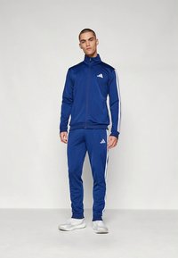 Blå mjukisdräkt i lättviktstyg med vita ränder längs ärmar och ben, jacka med dragkedja och logotyp på bröst och lår.