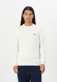 Hvit kabelstrikket genser med rund hals og ribbestrikkede mansjetter, med en liten grønn brodert logo på brystet.