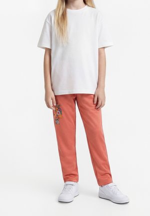 Enfant portant un t-shirt blanc uni à manches courtes, un pantalon corail avec broderie florale sur la cuisse gauche, et des baskets blanches, debout sur fond blanc.
