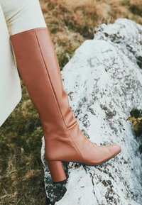 Botă de piele maro, până la genunchi, cu vârf ascuțit, toc bloc și textură netedă, așezată pe un fundal stâncos.