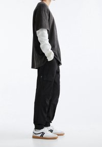 Schwarzes oversized T-Shirt über einem weißen Langarmshirt, kombiniert mit schwarzen Cargo-Hosen und weißen Sneakers mit schwarzen Akzenten.