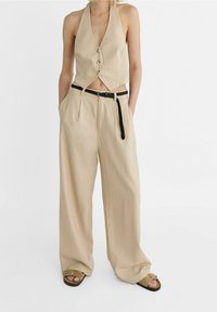 Beżowy krótki top na szyję z guzikami i wycięciem; w zestawie z luźnymi beżowymi spodniami i czarnym plecionym paskiem. Obuwie zdobione klamrą.