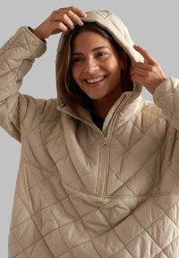 Beige quilted pullover hoodie with a half-zip front, diamond pattern, and a large hood. Textured fabric with a soft finish.