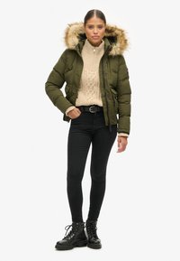 Olijfgroene gewatteerde jas met een bont gevoerde capuchon, gecombineerd met een beige kabelbrei truien, zwarte skinny jeans en zwarte veterschoenen.