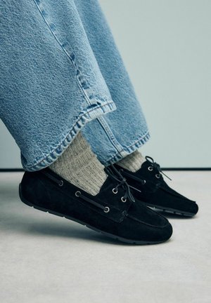 Scarpe in suede nere con lacci, dotate di occhielli in argento. Indossate con calze grigie a coste e pantaloni in denim blu con orlo sfilacciato.
