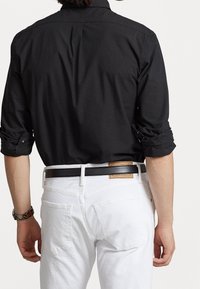 Maglietta nera a maniche lunghe con polsini arrotolati, abbinata a jeans di denim bianchi e una cintura nera. Presenta un'etichetta visibile sulla cintura.