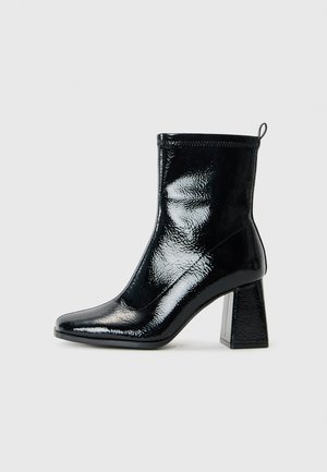 Bottine en patin noir avec un bout carré et un talon géométrique épais. Présente une texture lisse et brillante ainsi qu'une languette à l'arrière pour un enfilage facile.