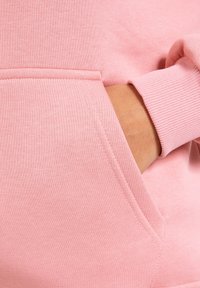 Sweatshirt rose avec une texture douce, doté d'une poche avant et de poignets côtelés. Le design présente des coutures nettes et un style minimaliste.