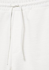 Gros plan de la taille d’un pantalon de jogging blanc avec bande élastique et longs cordons pendants au centre devant.