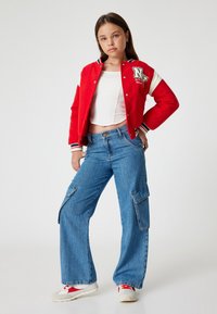 Chaqueta estilo varsity roja con mangas blancas y logo, combinada con pantalones cargo de denim azul de pierna ancha con bolsillos. Zapatillas blancas con detalles rojos.
