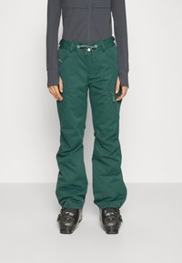 Groene skipants met een trekkoord in de taille, voorzakken en een ritszijzak, gecombineerd met zwarte skiboots en een nauwsluitend grijs shirt.