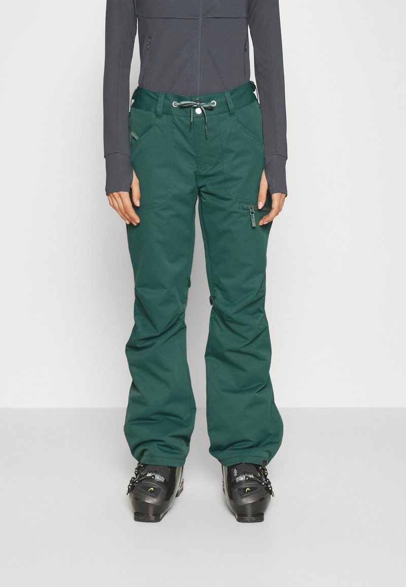 Groene skipants met een trekkoord in de taille, voorzakken en een ritszijzak, gecombineerd met zwarte skiboots en een nauwsluitend grijs shirt.