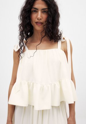 Haut à manches courtes en crème clair avec un ourlet volanté, des liens d'épaule ajustables et une texture douce. Design minimaliste avec une silhouette fluide.