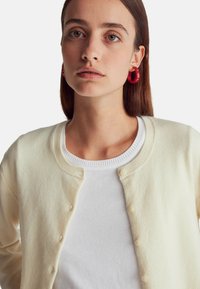 Kremowy kardigan z okrągłym dekoltem, z zapięciem na guziki i ściśniętymi mankietami, założony na białą koszulę z okrągłym dekoltem. Gładka tekstura, swobodny fason.