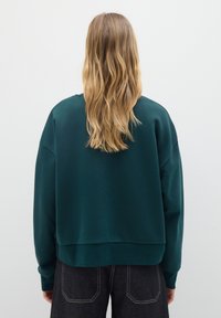 Sudadera recortada de color verde oscuro con cuello redondo y hombros caídos, que presenta puños y dobladillo acanalados. Llevada con jeans de mezclilla oscura.
